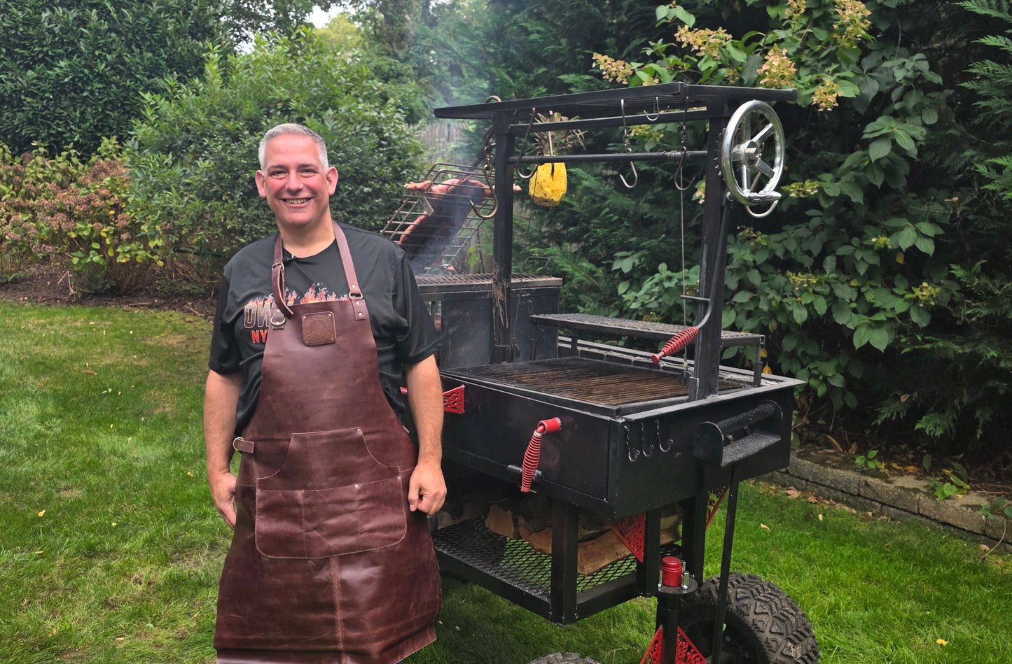 Mike Riddle wearing Artisan Leather apron standing with custom heavy gauge steel reverse flow smoker and Santa Maria grill in professional backyard cooking setup