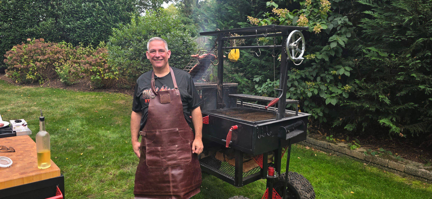 Mike Riddle wearing Gotham City Leather apron standing with custom heavy gauge steel reverse flow smoker and Santa Maria grill in professional backyard cooking setup