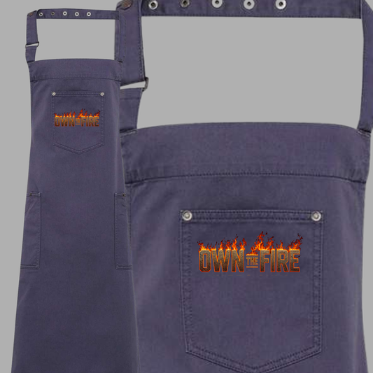 Navy blue apron with 'Own the Fire' text on a gray background
