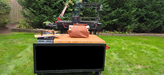 Own the Fire™ portable BBQ cart with Gotham City Leather split carrier and custom Santa Maria grill in professional backyard setup showing complete cooking ecosystem with Crafted in Smoke Forged in Fire branding